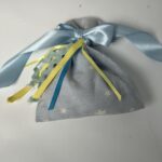 Αυτό το χειροποίητο πουγκί, φτιαγμένο από απαλό γαλάζιο ύφασμα με διακριτικά αστεράκια, είναι ένα μικρό έργο τέχνης. Η γαλάζια σατέν κορδέλα, δεμένη σε έναν κομψό φιόγκο, προσδίδει μια αίσθηση γιορτής, ενώ οι πολύχρωμες κορδέλες που ξεπροβάλλουν από το άνοιγμα του πουγκιού προσθέτουν μια παιχνιδιάρικη πινελιά. Το πουγκί, γεμάτο με άρωμα λεβάντας ή με μικρά δωράκια, είναι ιδανικό για να εκφράσει την αγάπη και την τρυφερότητα προς τους αγαπημένους σας.