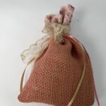 Αυτό το χειροποίητο πουγκί, φτιαγμένο από φυσική λινάτσα σε μια ζεστή ροζ απόχρωση, αποπνέει μια rustic γοητεία. Η λεπτή δαντέλα και οι σατέν κορδέλες, σε χρυσές και ροζ αποχρώσεις, προσθέτουν μια ρομαντική πινελιά. Το πουγκί, γεμάτο με αποξηραμένα λουλούδια ή αρωματικά βότανα, είναι ιδανικό για να χαρίσει μια αίσθηση ζεστασιάς και φροντίδας στους αγαπημένους σας.