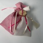 Μια ξεχωριστή μπομπονιέρα βάπτισης σε διχρωμία ροζ και ιβουάρ, κατασκευασμένη από υφασμάτινο τούλι με ανάλαφρη υφή. Είναι διακοσμημένη με λεπτές σατέν κορδέλες σε ροζ και λευκές αποχρώσεις, καθώς και ένα ξύλινο στοιχείο με χαραγμένο όνομα, που την καθιστά ιδιαίτερα προσωποποιημένη. Ένα μικρό διακριτικό φουντάκι ολοκληρώνει το κομψό σχέδιο, προσθέτοντας μια πινελιά πολυτέλειας.