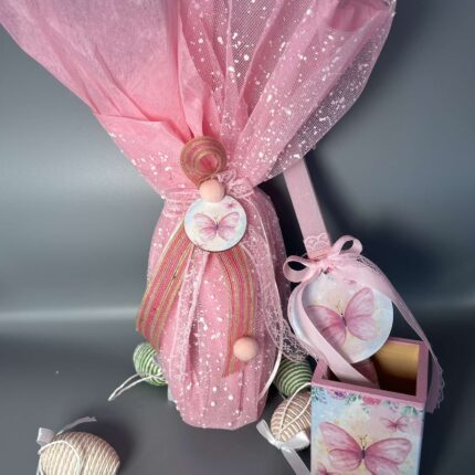 Αυτό το χειροποίητο πασχαλινό δώρο αποτελείται από μια λαμπάδα και μια μολυβοθήκη, που συνδυάζουν τη γιορτινή παράδοση με μια παιχνιδιάρικη διάθεση. Η λαμπάδα είναι τυλιγμένη με ροζ τούλι, διακοσμημένο με ασημί glitter, που προσδίδει μια αίσθηση λάμψης και μαγείας. Ένα μικρό ξύλινο διακοσμητικό με μια πεταλούδα στο κέντρο, στολισμένο με ροζ κορδέλες και pom poms, προσθέτει μια παιχνιδιάρικη πινελιά. Η μολυβοθήκη, σε ροζ αποχρώσεις, είναι διακοσμημένη με μια ζωγραφισμένη πεταλούδα, που ταιριάζει με το θέμα της λαμπάδας. Ένα μικρό στρογγυλό διακοσμητικό, επίσης με μια πεταλούδα, κρέμεται από τη μολυβοθήκη, προσθέτοντας μια έξτρα πινελιά ομορφιάς. Το δώρο πλαισιώνεται από μικρά, σχοινένια πασχαλινά αυγά σε φυσικές αποχρώσεις, δημιουργώντας μια αρμονική σύνθεση. Είναι ιδανικό για κορίτσια που αγαπούν τις πεταλούδες, τα χρώματα και τις χειροποίητες δημιουργίες.