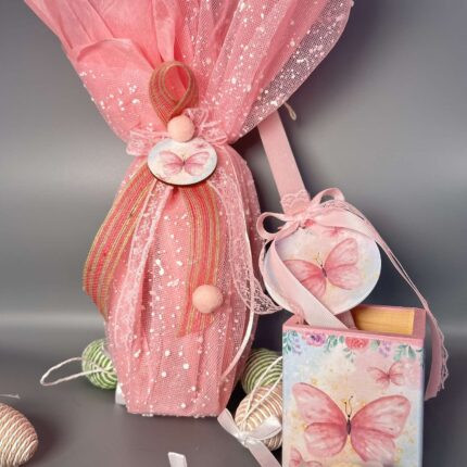 Αυτό το χειροποίητο πασχαλινό δώρο αποτελείται από μια λαμπάδα και μια μολυβοθήκη, που συνδυάζουν τη γιορτινή παράδοση με μια παιχνιδιάρικη διάθεση. Η λαμπάδα είναι τυλιγμένη με ροζ τούλι, διακοσμημένο με ασημί glitter, που προσδίδει μια αίσθηση λάμψης και μαγείας. Ένα μικρό ξύλινο διακοσμητικό με μια πεταλούδα στο κέντρο, στολισμένο με ροζ κορδέλες και pom poms, προσθέτει μια παιχνιδιάρικη πινελιά. Η μολυβοθήκη, σε ροζ αποχρώσεις, είναι διακοσμημένη με μια ζωγραφισμένη πεταλούδα, που ταιριάζει με το θέμα της λαμπάδας. Ένα μικρό στρογγυλό διακοσμητικό, επίσης με μια πεταλούδα, κρέμεται από τη μολυβοθήκη, προσθέτοντας μια έξτρα πινελιά ομορφιάς. Το δώρο πλαισιώνεται από μικρά, σχοινένια πασχαλινά αυγά σε φυσικές αποχρώσεις, δημιουργώντας μια αρμονική σύνθεση. Είναι ιδανικό για κορίτσια που αγαπούν τις πεταλούδες, τα χρώματα και τις χειροποίητες δημιουργίες.