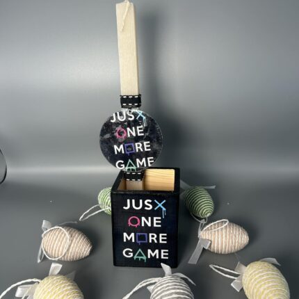 Ένα ξεχωριστό πασχαλινό σετ για λάτρεις του gaming! Περιλαμβάνει μια ξύλινη λαμπάδα και μια μολυβοθήκη σε μαύρο χρώμα, διακοσμημένες με το εμβληματικό σύνθημα "Just One More Game". Η λαμπάδα και η μολυβοθήκη πωλούνται και ξεχωριστά.