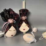 Αυτό το χειροποίητο πασχαλινό δώρο αποτελείται από μια αυγούλα και μια λαμπάδα, που ξεχωρίζουν για την καλλιτεχνική τους αισθητική. Και τα δύο αντικείμενα είναι διακοσμημένα με ζωγραφισμένα πρόσωπα με κλειστά μάτια και κόκκινα χείλη, δημιουργώντας μια αίσθηση ηρεμίας και ομορφιάς. Είναι ντυμένα με κομψά υφάσματα σε σκούρες αποχρώσεις με φλοράλ μοτίβα, που προσδίδουν μια αίσθηση πολυτέλειας και μυστηρίου. Η αυγούλα είναι στολισμένη με μια ροζ τριαντάφυλλο και μια πέρλα, ενώ η λαμπάδα με ένα απλό ροζ τριαντάφυλλο. Είναι ιδανικά για όσους εκτιμούν την τέχνη, την κομψότητα και τις χειροποίητες δημιουργίες.