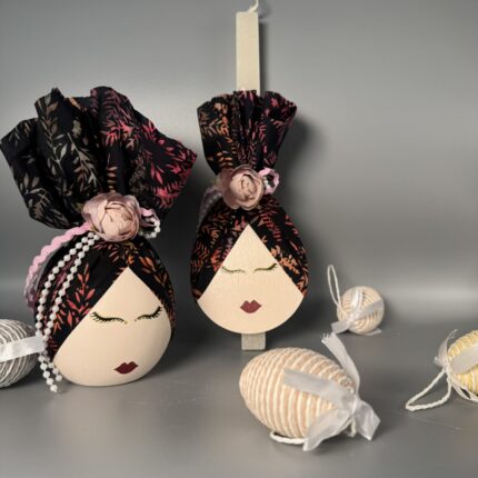 Αυτό το χειροποίητο πασχαλινό δώρο αποτελείται από μια αυγούλα και μια λαμπάδα, που ξεχωρίζουν για την καλλιτεχνική τους αισθητική. Και τα δύο αντικείμενα είναι διακοσμημένα με ζωγραφισμένα πρόσωπα με κλειστά μάτια και κόκκινα χείλη, δημιουργώντας μια αίσθηση ηρεμίας και ομορφιάς. Είναι ντυμένα με κομψά υφάσματα σε σκούρες αποχρώσεις με φλοράλ μοτίβα, που προσδίδουν μια αίσθηση πολυτέλειας και μυστηρίου. Η αυγούλα είναι στολισμένη με μια ροζ τριαντάφυλλο και μια πέρλα, ενώ η λαμπάδα με ένα απλό ροζ τριαντάφυλλο. Είναι ιδανικά για όσους εκτιμούν την τέχνη, την κομψότητα και τις χειροποίητες δημιουργίες.