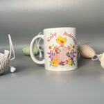 Αυτή η λευκή κούπα δεν είναι απλώς ένα καθημερινό αντικείμενο, αλλά ένα μικρό έργο τέχνης που φέρνει την ατμόσφαιρα του Πάσχα και της άνοιξης απευθείας στο χέρι σου. Στο κέντρο της, μια χαριτωμένη πεταλούδα με ζωηρά χρώματα ξεδιπλώνει τα φτερά της, ενώ ένα γλυκό μελισσάκι και ένα νεοσσός που μόλις βγήκε από το αυγό προσθέτουν μια παιχνιδιάρικη πινελιά. Τα μικρά πράσινα φύλλα που σκορπίζονται γύρω τους ενισχύουν την αίσθηση της αναγέννησης και της φύσης που ανθίζει. Η χειρόγραφη επιγραφή "Καλή Ανάσταση!" με τα μοβ γράμματα προσθέτει μια προσωπική και ζεστή ευχή. Η κούπα περιβάλλεται από πασχαλινά αυγά από σπάγγο σε απαλές αποχρώσεις, δημιουργώντας μια αίσθηση γιορτής και αρμονίας. Αυτή η κούπα είναι το ιδανικό δώρο για να χαρίσετε χαρά και αισιοδοξία σε αγαπημένα σας πρόσωπα, φέρνοντας το πνεύμα του Πάσχα στην καθημερινότητά τους.